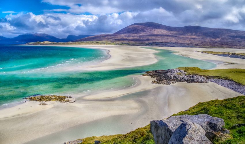 A series of 2 striking images of the beaches at Luskentyre on Eilean Siar in the Western Isles or Outer Hebrides (The Isle of Harris, specifically), off North West Scotland in the UK. Taken in midmorning, early May 2021 during an unusually prolonged winter. Both images show dramatic storm clouds building over the North Harris hills but these are casing shadows and unusual light where the hills meet the sea. The sand and sea at this world famous viewpoint are constantly shifting such that a new sandbank has developed and allowed the Jade Green seas to create new routes over the dazzling white sand. The two pictures are broadly similar but the second one is arguably more dramatic, given the shooting in HDR. In the first picture illustrates the deep cobalt blue sea, fading towards the hills as deep emerald but then fades further still to paler Jade in the shallowest areas. The second picture was taken in HDR and really emphasises the already vivid colours of the water, the rocks and the sand.